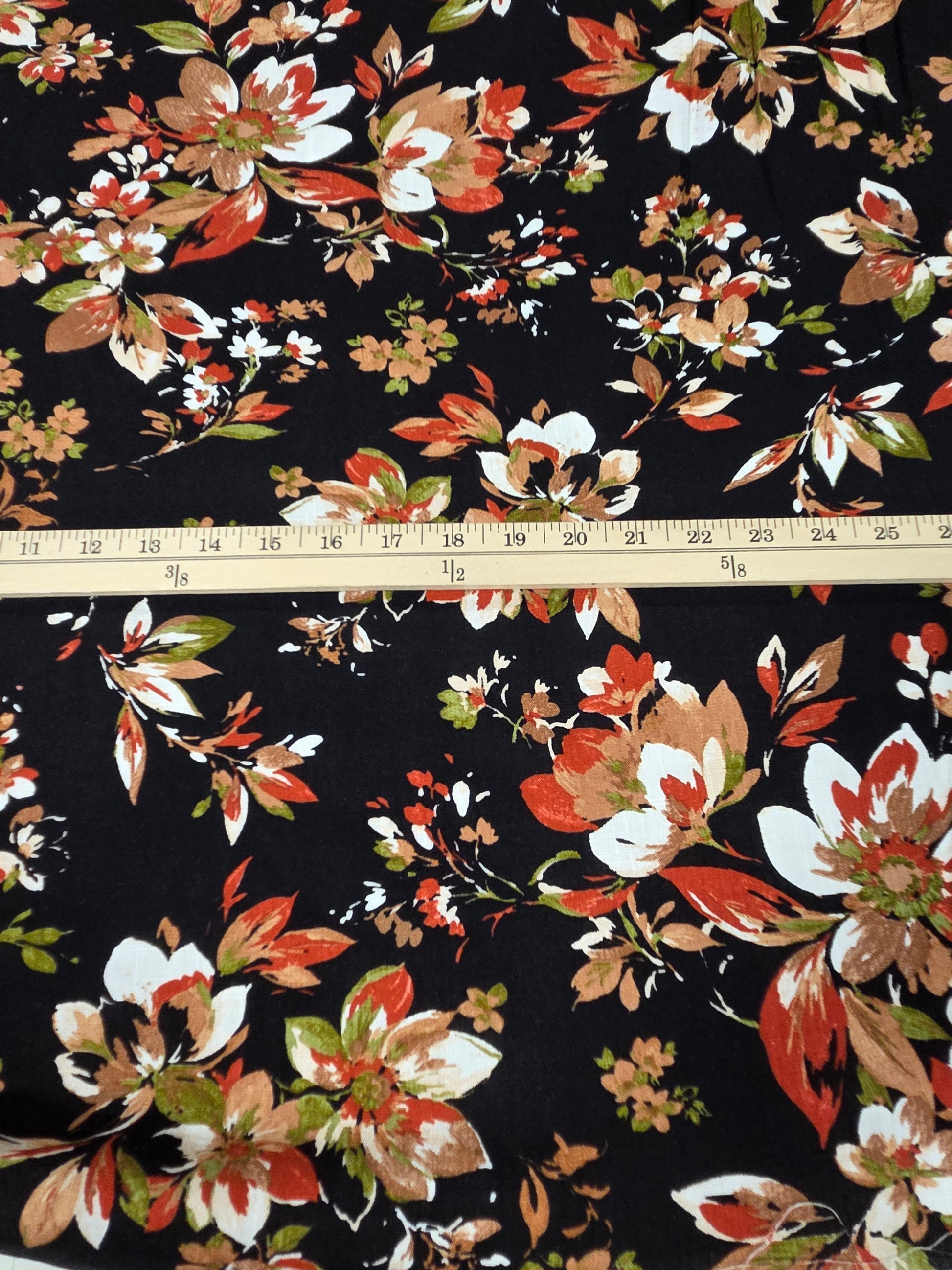 Black floral fabric with a repeating pattern of white, orange, and brown flowers, shown with a wooden ruler across the middle for scale.