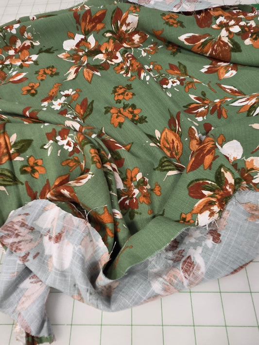A piece of green fabric with a brown, orange, and white floral print, layered with a sheer light gray plaid lining, laid out on a white cutting mat.