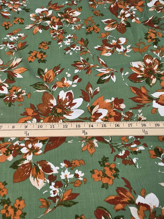 A piece of green fabric with a vintage floral print in orange, brown, and white, laid flat on a cutting mat with a wooden ruler showing its width.