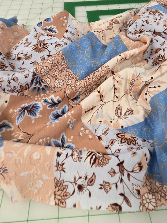 A close-up of a patchwork fabric with floral and botanical prints in beige, brown, blue, and white, laid out on a green grid cutting mat with a wooden ruler visible in the background.