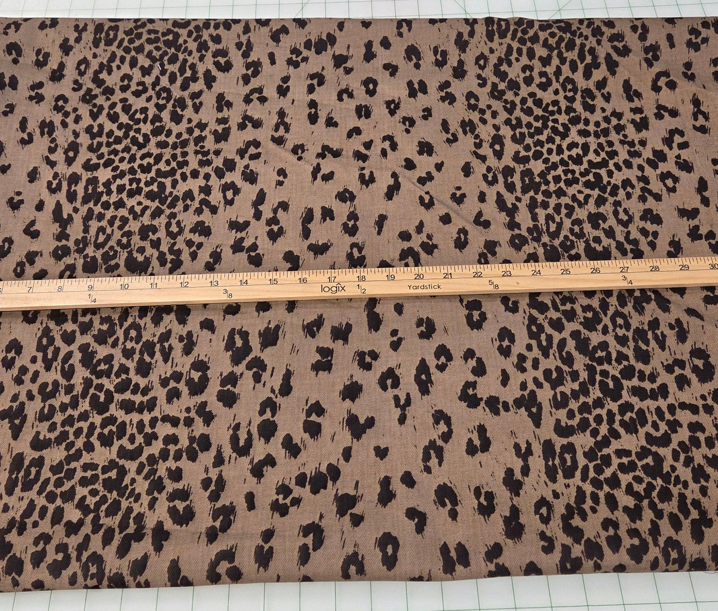 A large rectangular piece of leopard print fabric in brown and black is laid flat on a white measuring mat with a wooden ruler across the middle, with other folded fabrics visible in the background.