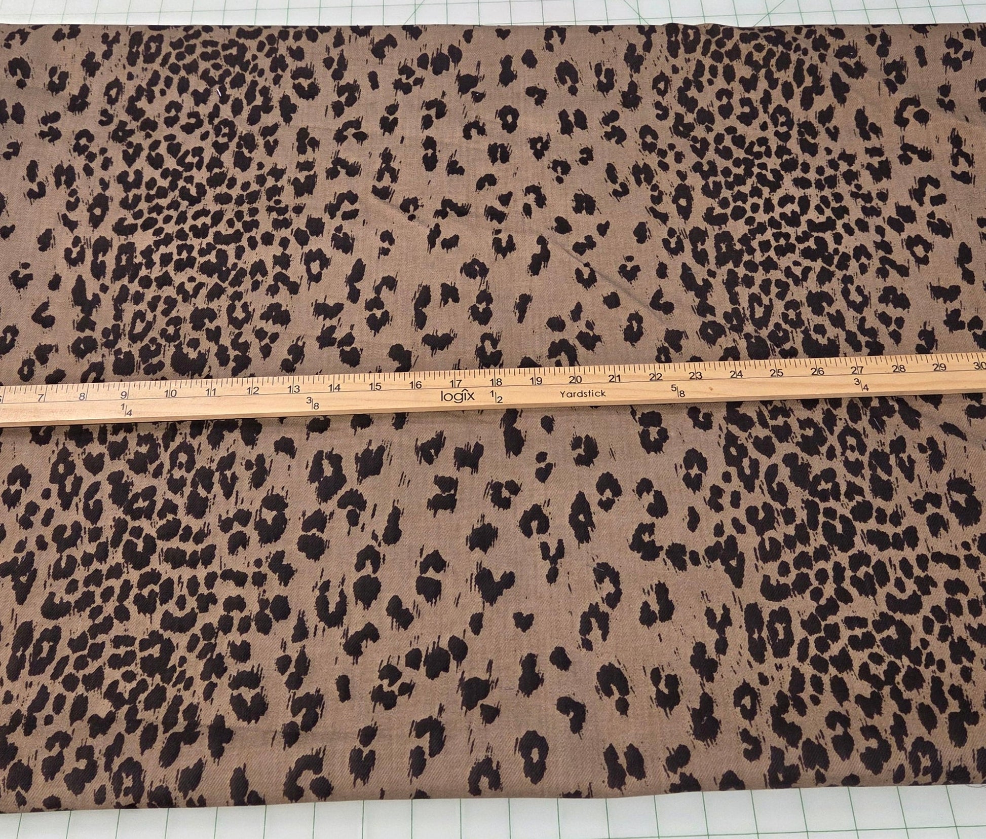 A large rectangular piece of leopard print fabric in brown and black is laid flat on a white measuring mat with a wooden ruler across the middle, with other folded fabrics visible in the background.