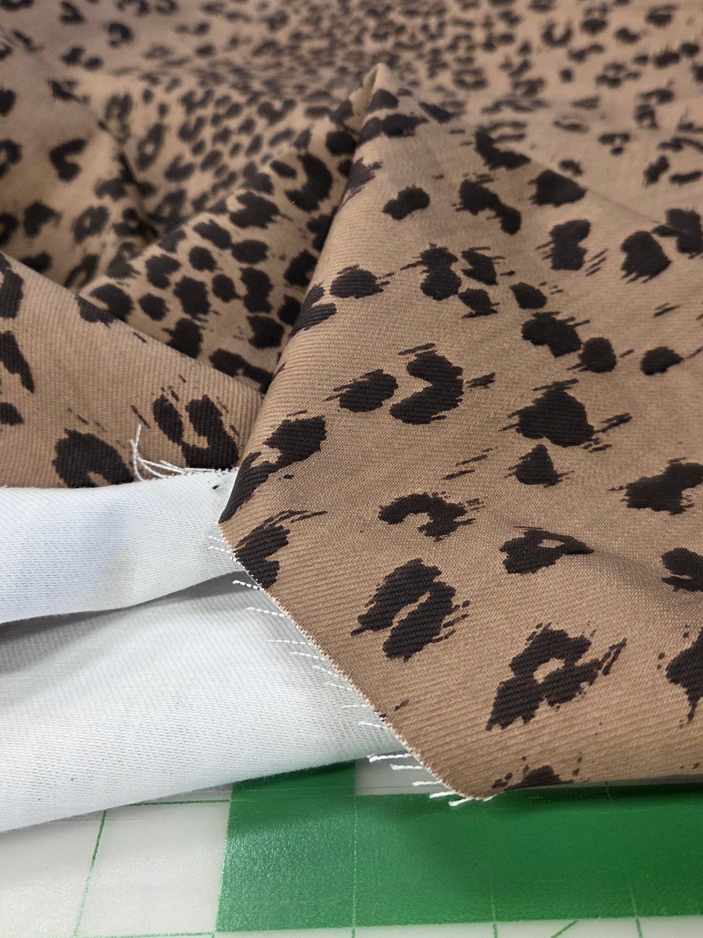 Brown and black leopard print fabric draped over a white fabric piece on a green cutting mat, with other fabrics visible in the background.