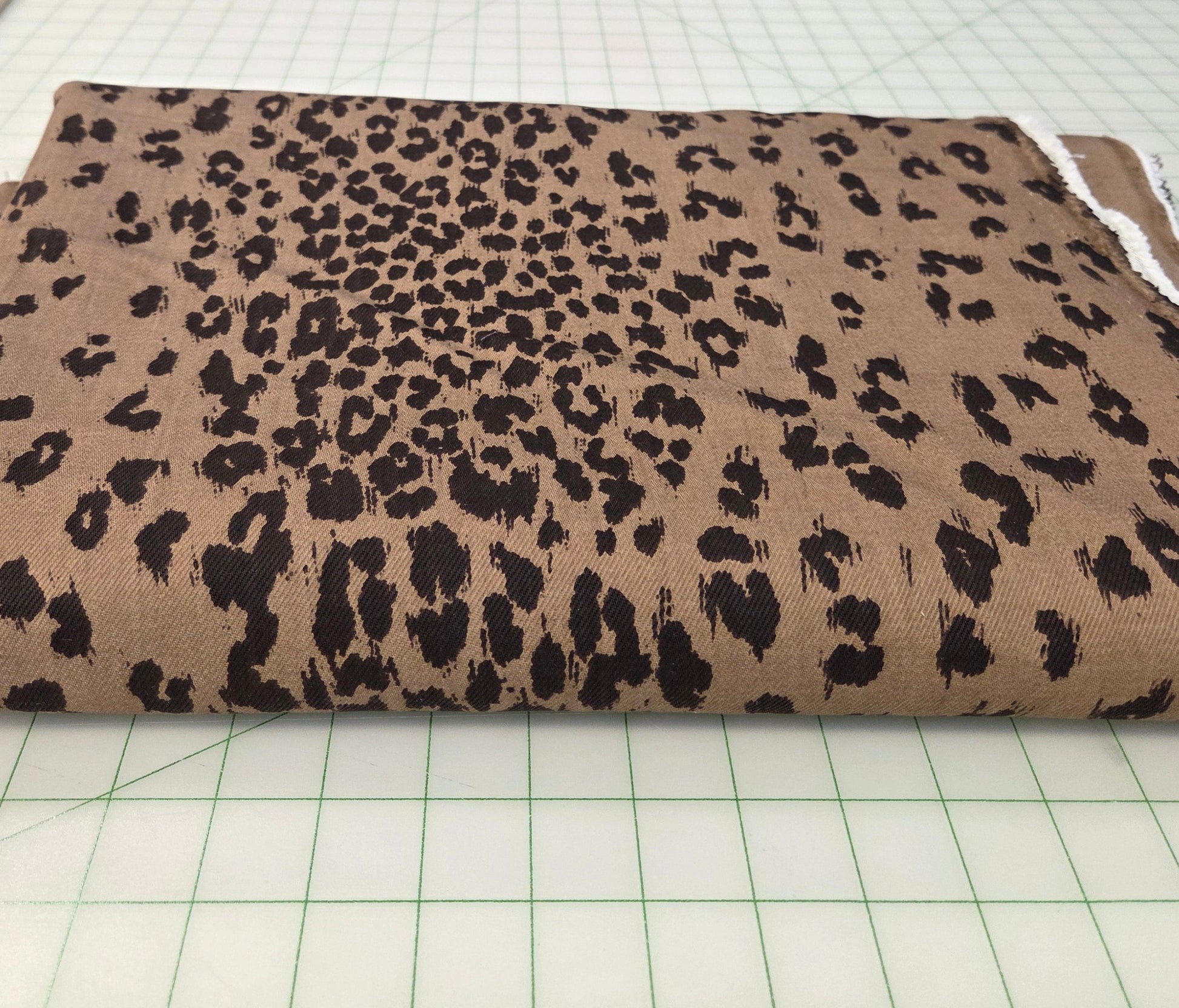 A folded piece of leopard print fabric with black spots on a tan background, displayed on a green grid cutting mat in a fabric store.