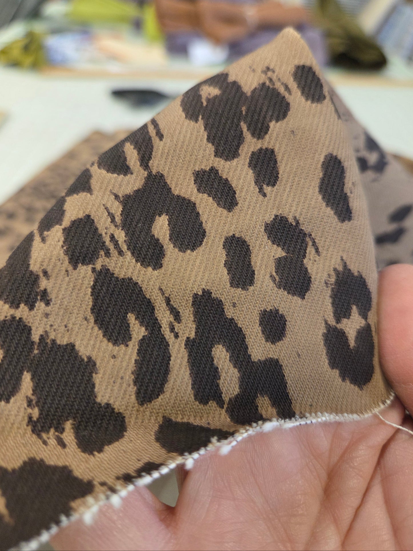 A close-up of a hand holding a triangular piece of leopard print fabric with a tan background and dark brown spots, showing the raw woven edge.