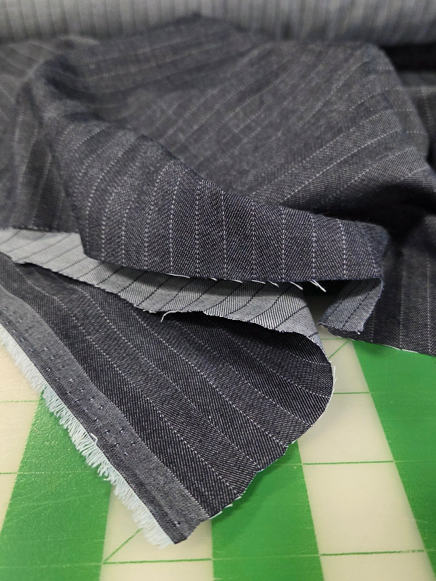 A close-up of dark gray pinstriped fabric draped over a green cutting mat, with a matching gray fabric roll visible in the background.