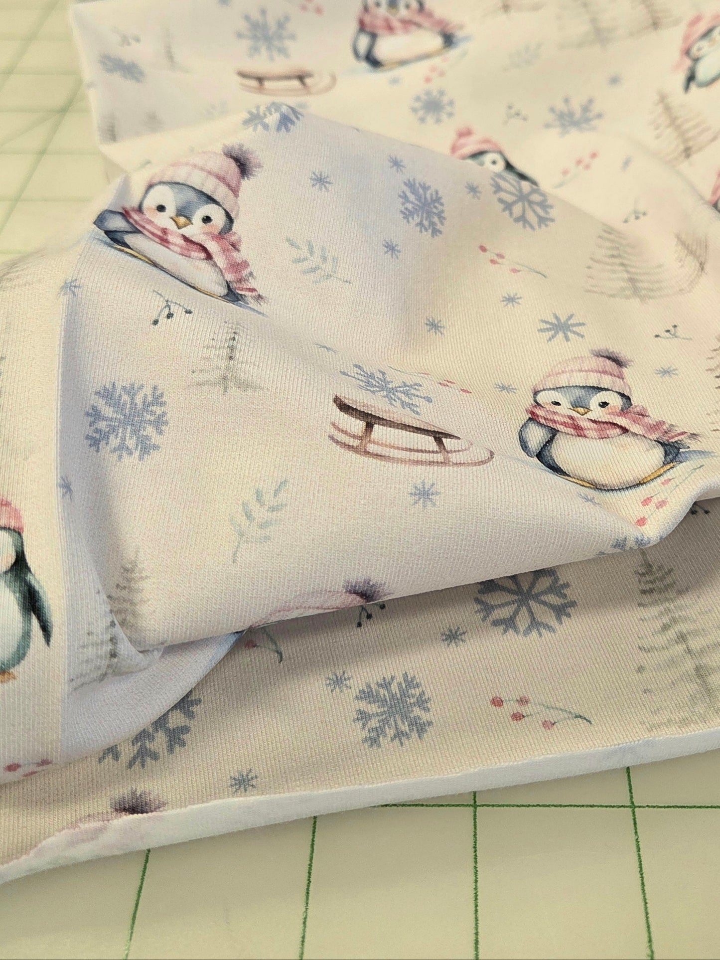 A close-up of a fabric yardage featuring a winter penguin print on a cream background, with penguins wearing pink and blue scarves and hats, blue snowflakes, and small evergreen trees. The fabric is draped over a green cutting mat.