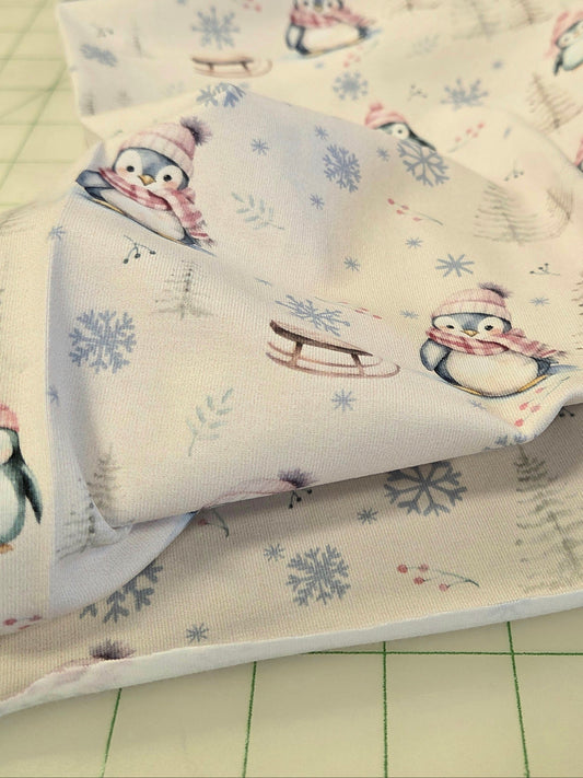 A close-up of a fabric yardage featuring a winter penguin print on a cream background, with penguins wearing pink and blue scarves and hats, blue snowflakes, and small evergreen trees. The fabric is draped over a green cutting mat.