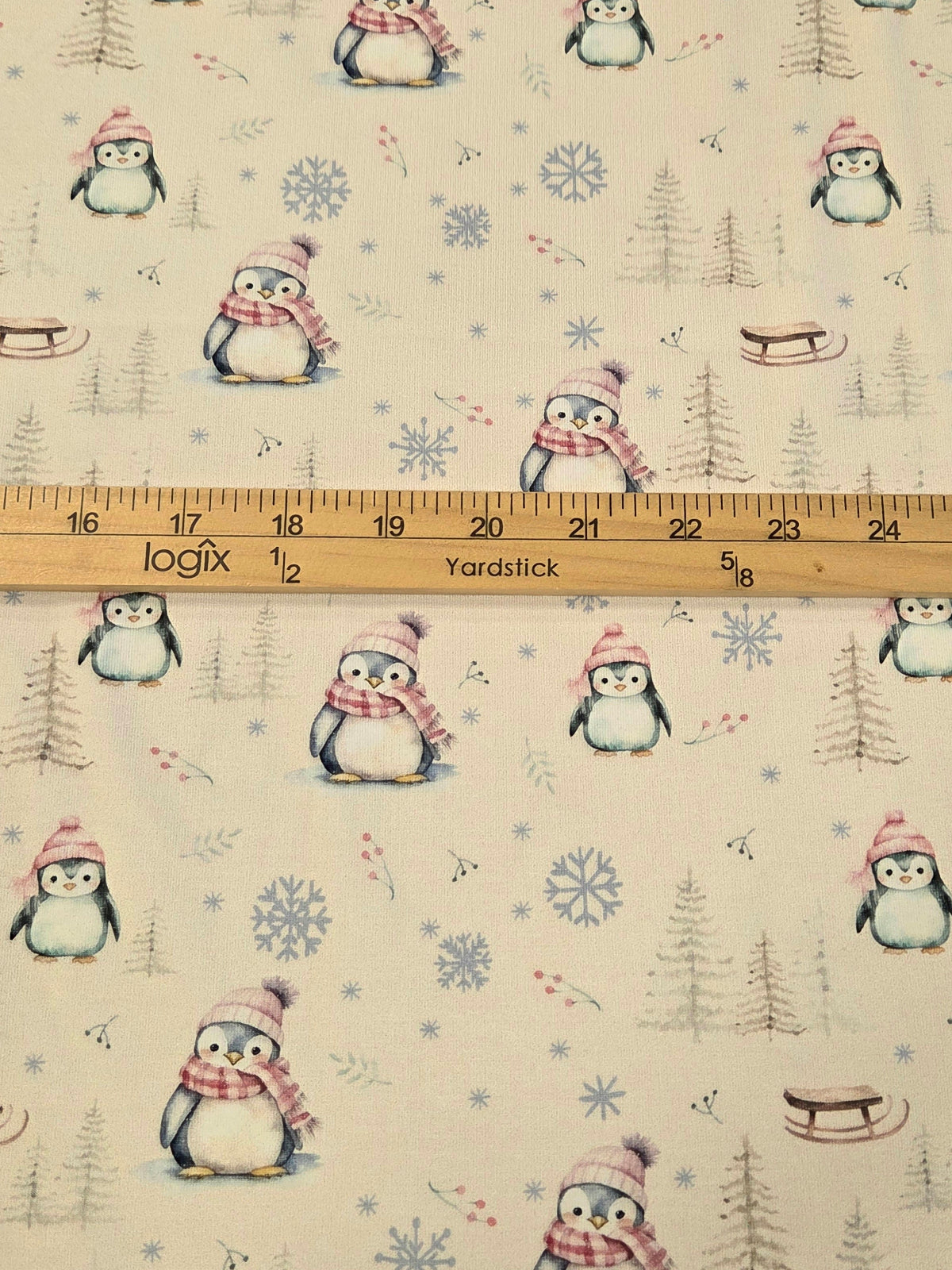 A close-up of a fabric pattern featuring penguins wearing pink and red scarves, set against a light cream background with blue snowflakes and gray evergreen trees. A wooden yardstick is placed horizontally across the center to show the scale of the pattern.