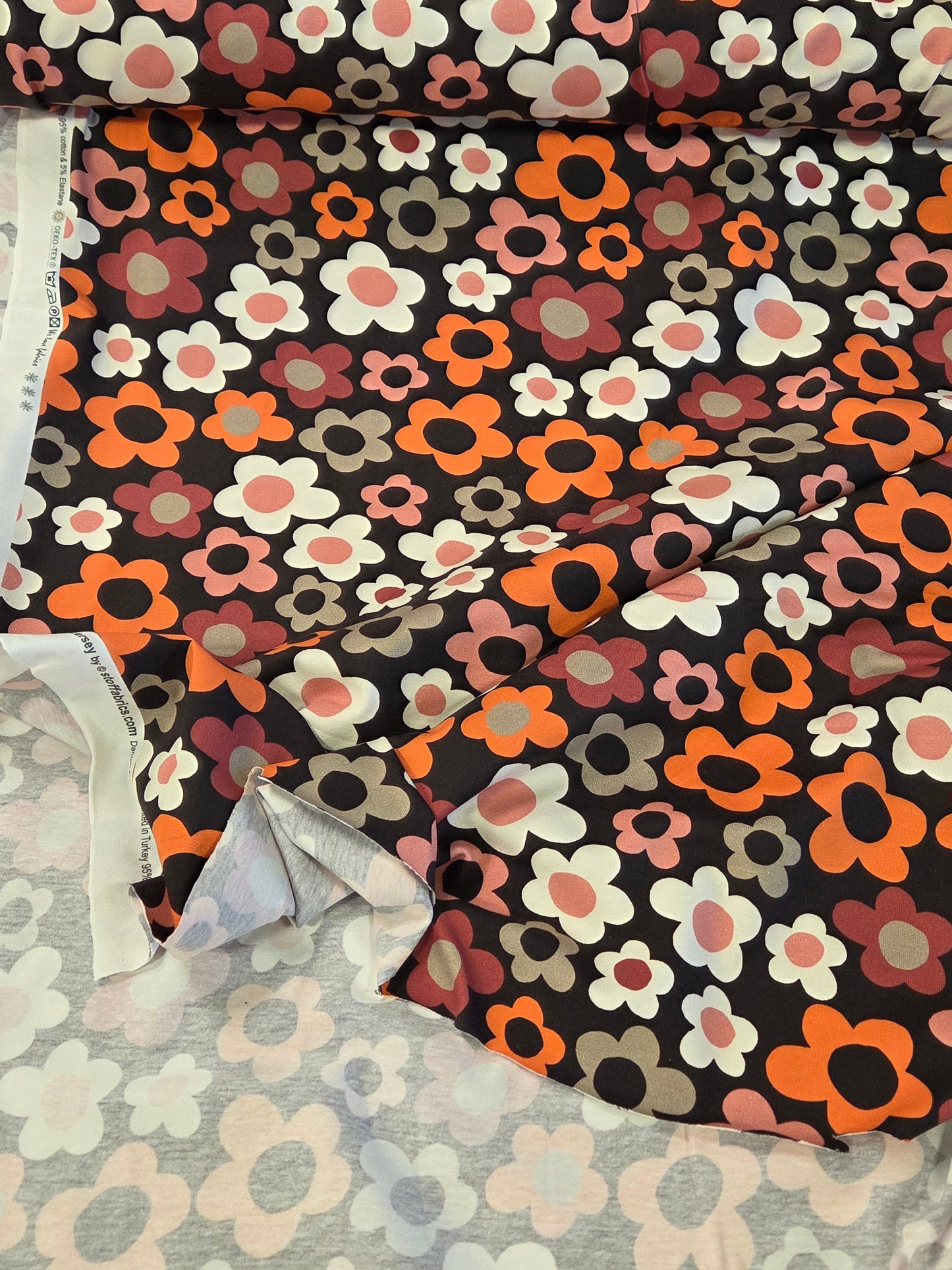 A roll of floral fabric with a retro pattern of orange, pink, white, and gray flowers on a black background, laid out next to another patterned fabric. Stof fabrics OEKO tex