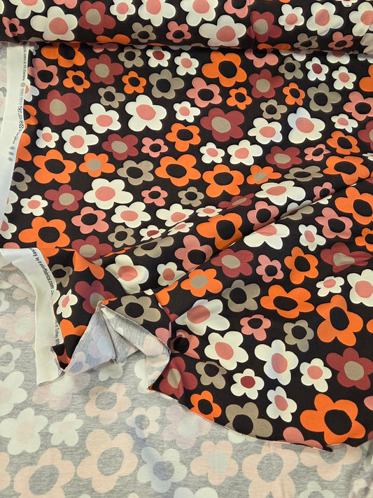 A roll of floral fabric with a retro pattern of orange, pink, white, and gray flowers on a black background, laid out next to another patterned fabric. Stof fabrics OEKO tex