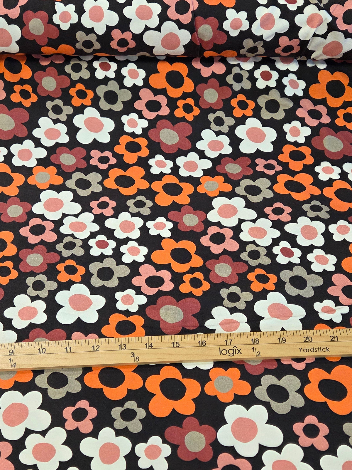 A roll of floral fabric with a retro 1970s pattern, shown alongside a wooden ruler for scale. The fabric features orange, pink, white, and gray flowers on a black background.