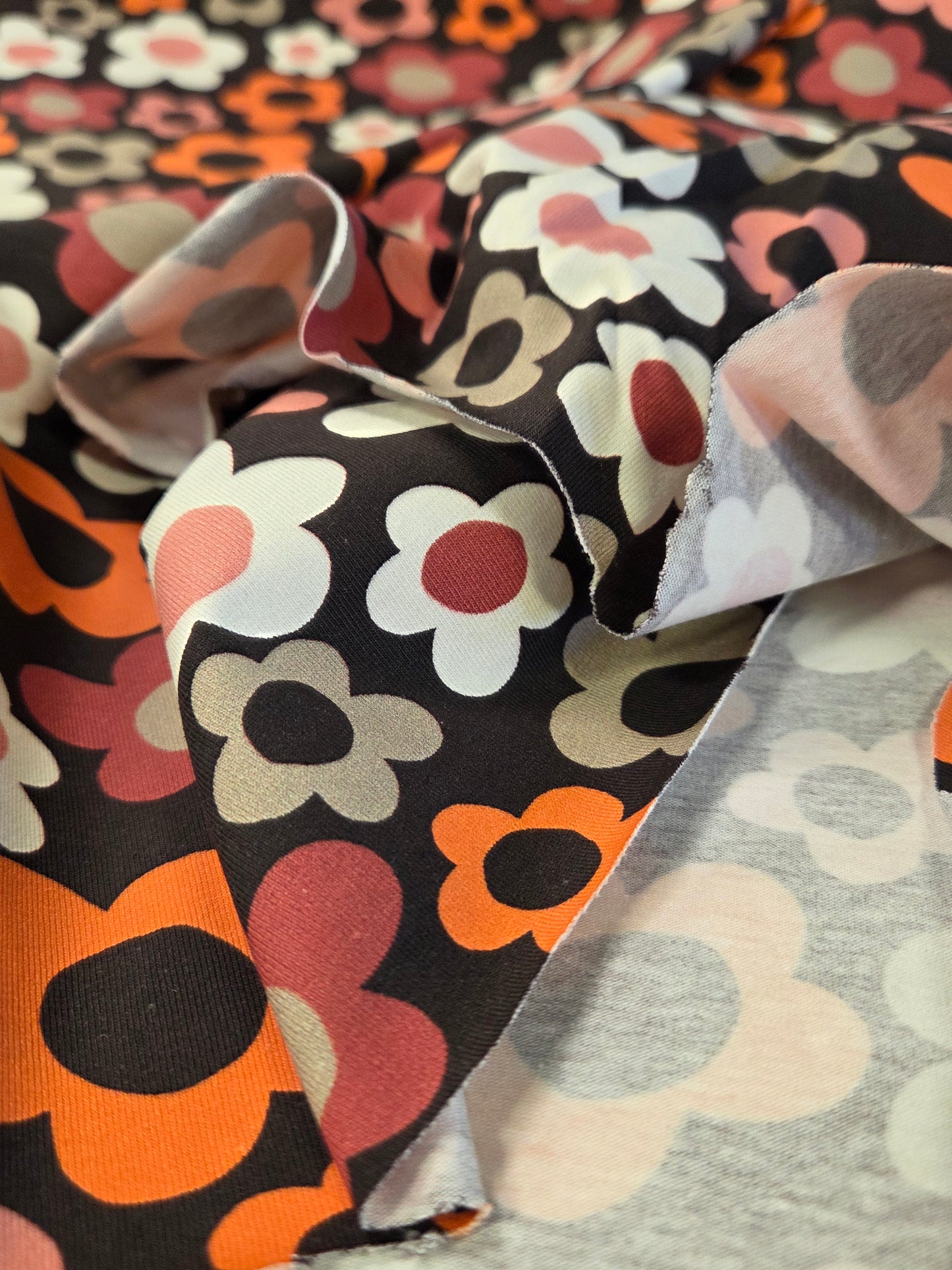 A close-up of patterned fabric with a retro floral print. The fabric features large stylized flowers in orange, pink, white, and gray on a black background, with a lighter gray fabric layered underneath. Stof Fabrics. OEKO TEX certified