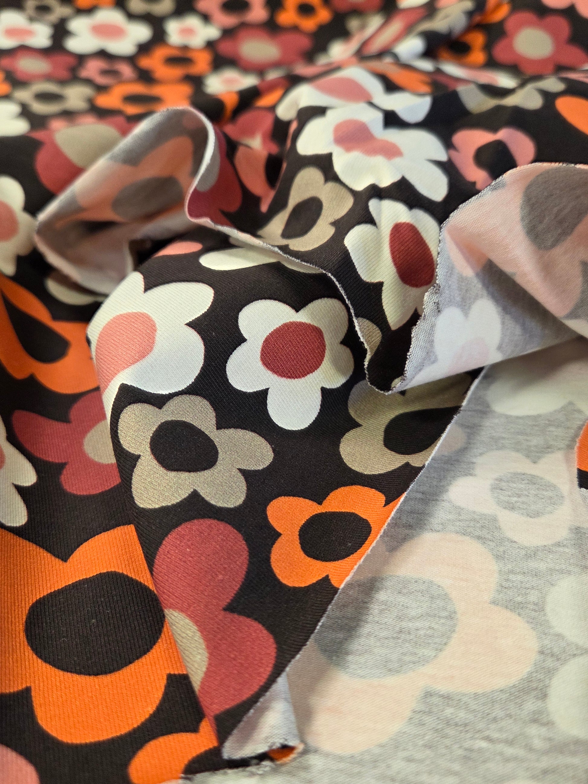 A close-up of patterned fabric with a retro floral print. The fabric features large stylized flowers in orange, pink, white, and gray on a black background, with a lighter gray fabric layered underneath. Stof Fabrics. OEKO TEX certified