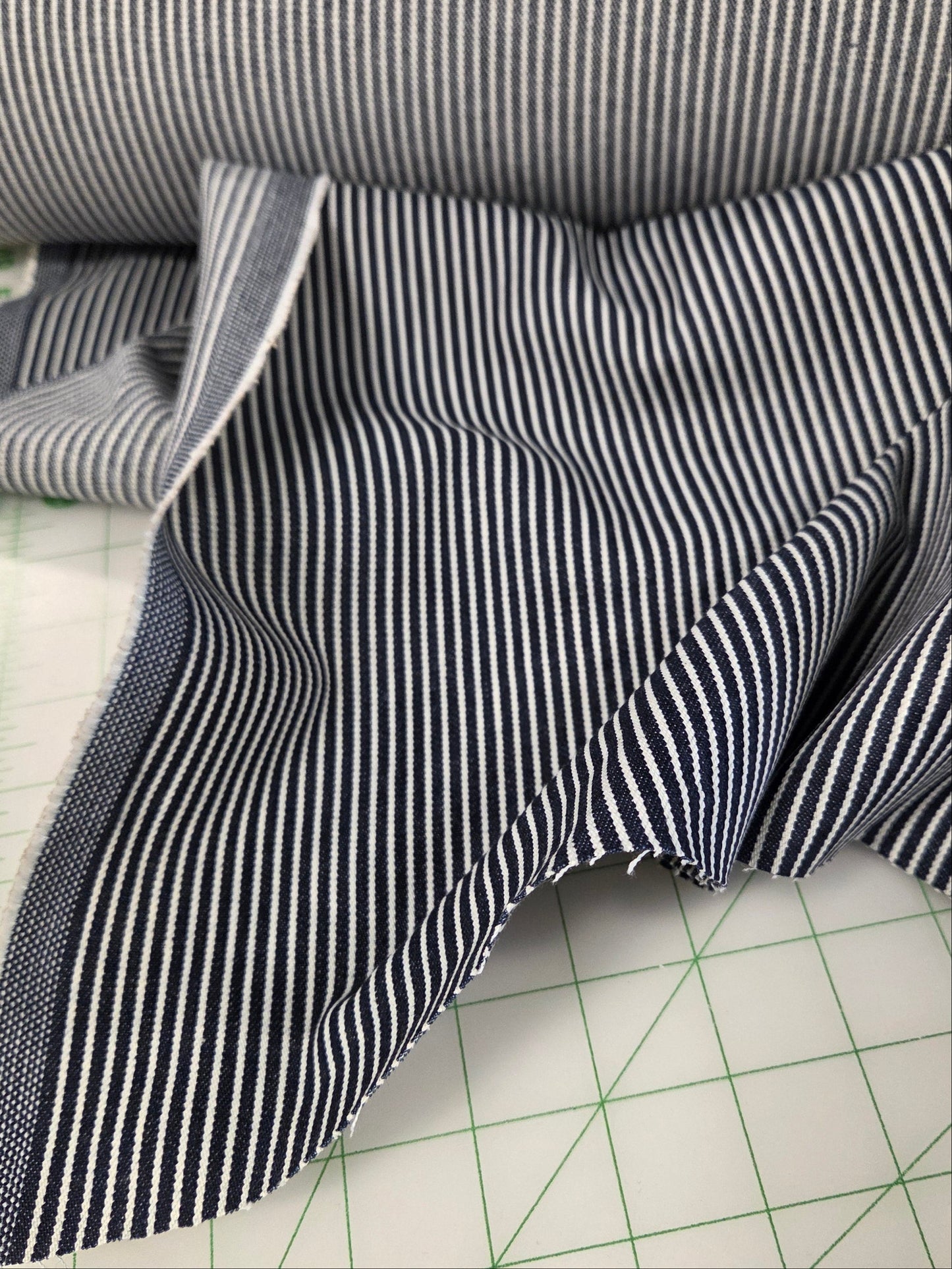 A close-up of striped fabric, with a roll at the top and a folded piece spread out on a green grid cutting mat. The fabric has thin dark stripes on a light gray or off-white base.