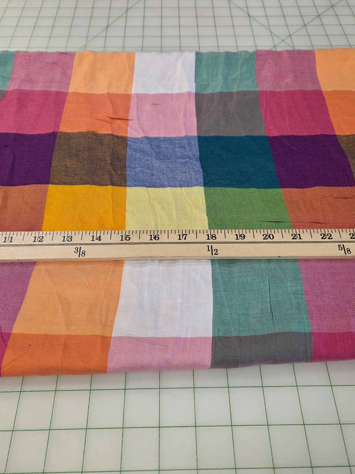A folded piece of multicolor patchwork fabric with a grid pattern of colored squares, shown on a green gridded cutting mat with a wooden ruler for scale.