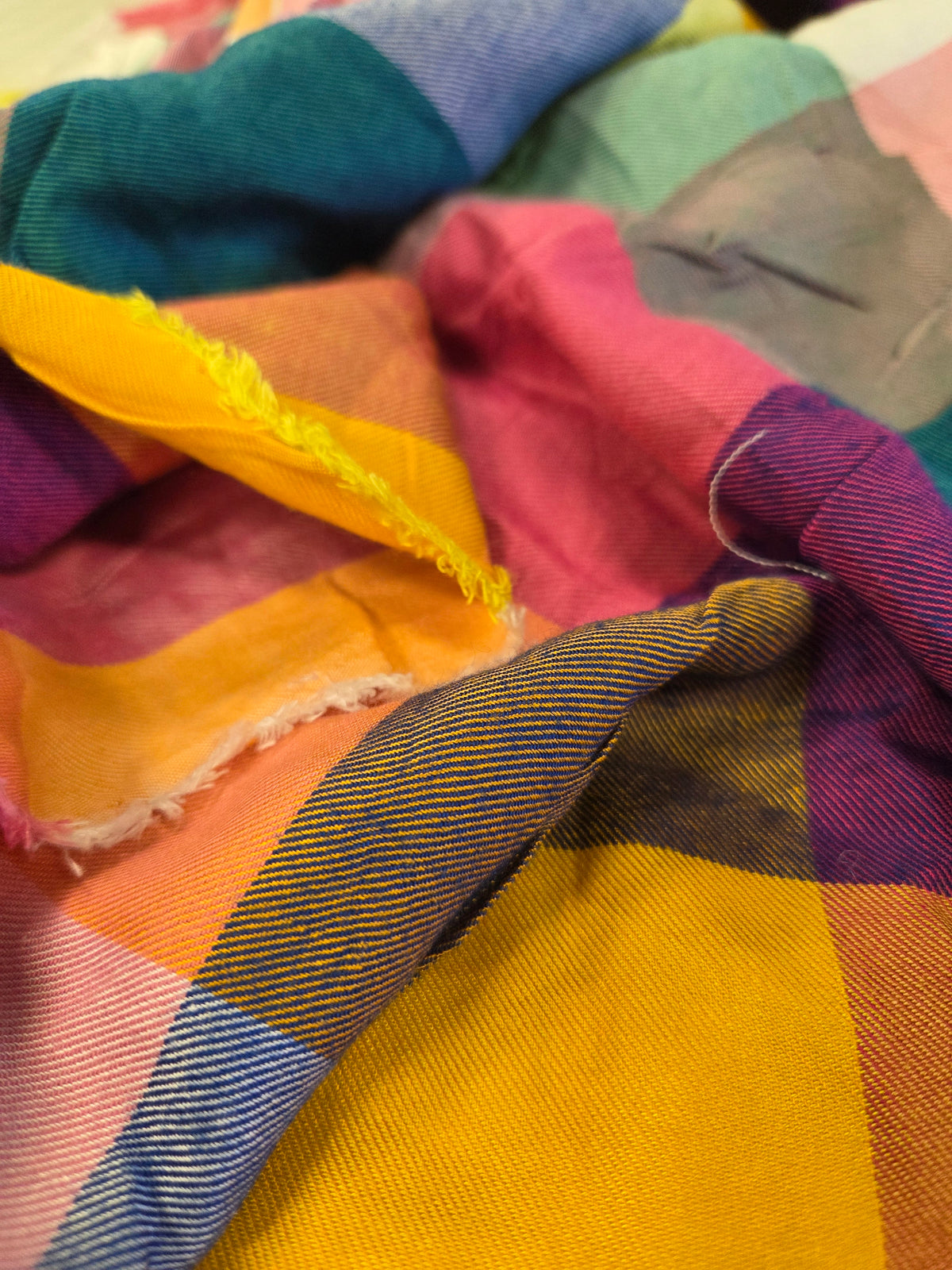 A close-up of a multicolor plaid check fabric with a mix of bright colors including yellow, pink, blue, purple, and orange, showing its woven texture and frayed edges. LA Finch Fabrics retail by the yard online shipping to US