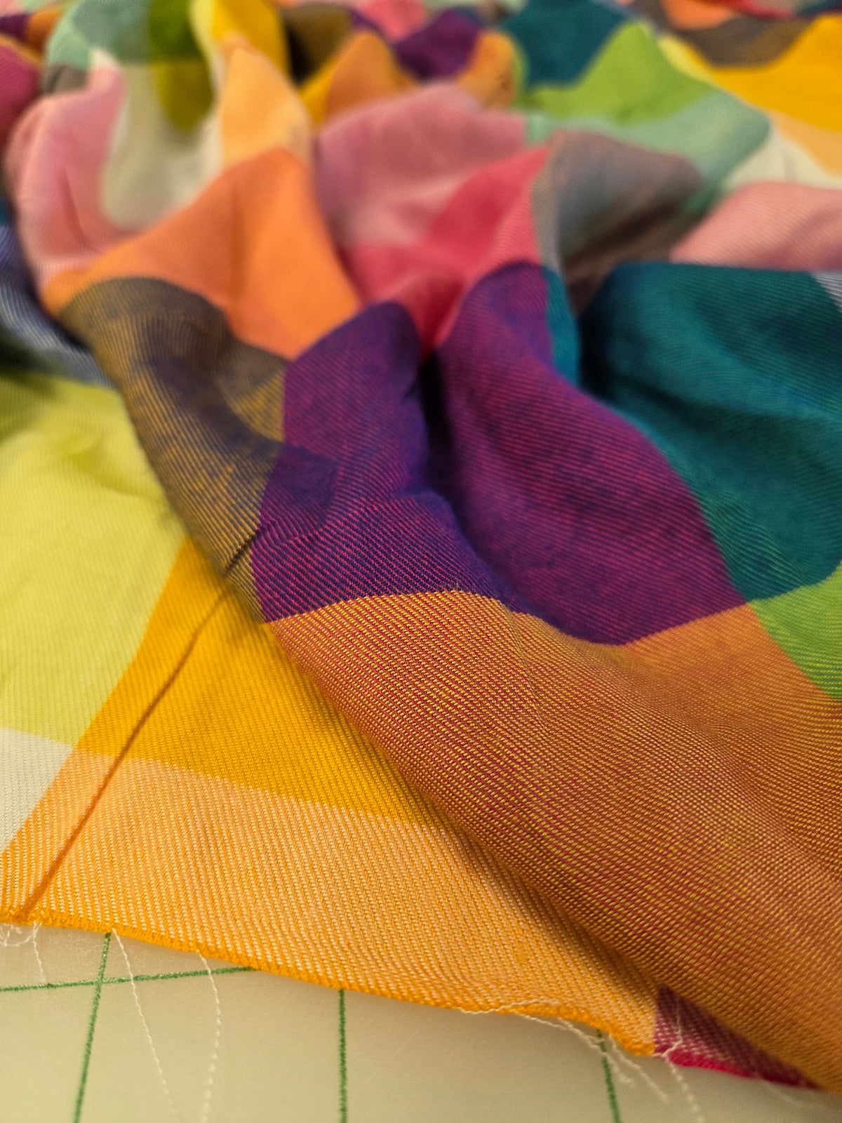 A close-up of a multicolor woven fabric with large blocks of yellow, orange, purple, teal, pink, green, and white, draped over a light-colored surface with visible thread and grid lines.