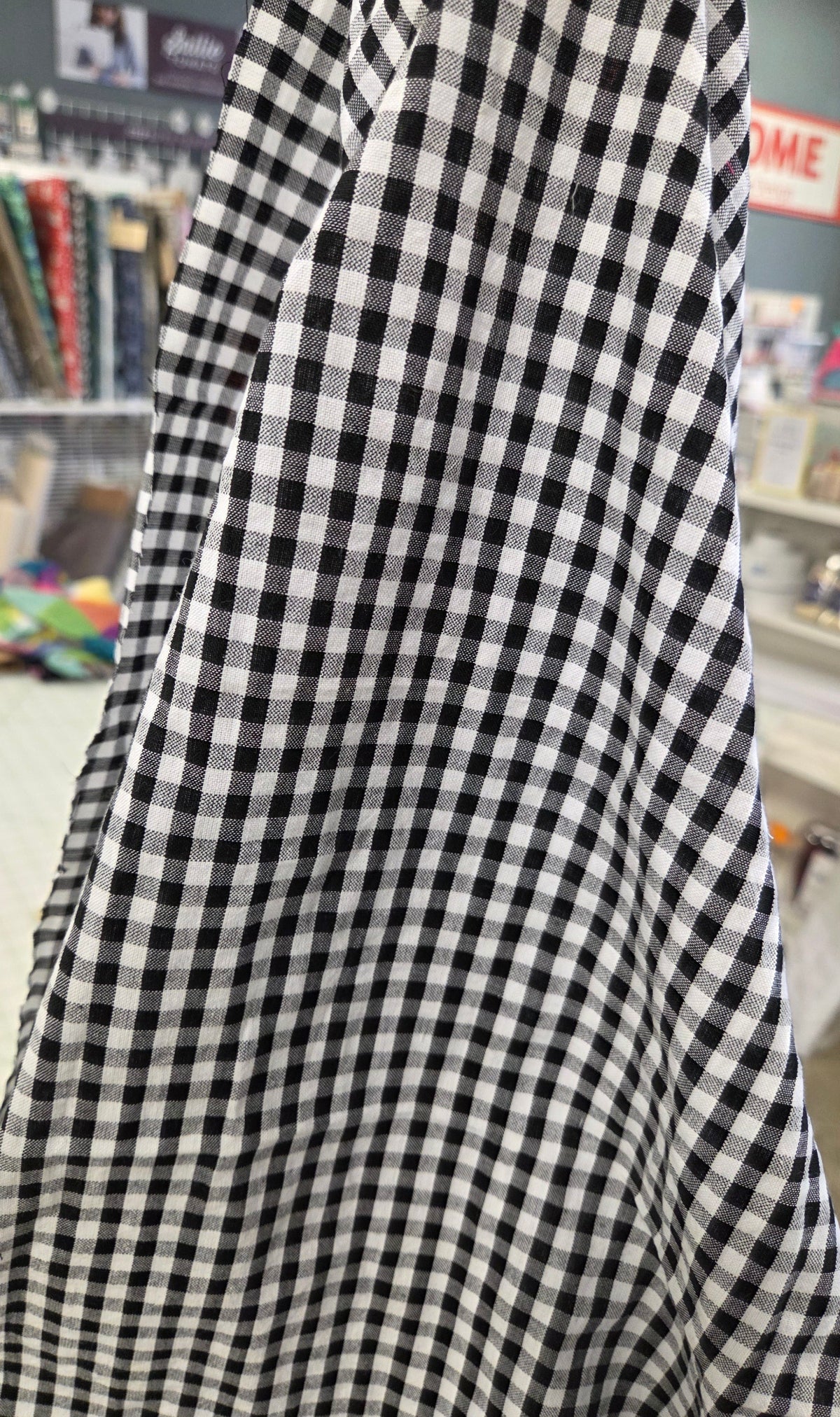 A close-up of a black and white gingham fabric draped vertically, with a blurred store interior in the background.  Apparel Fabric Shop Cotton Gingham