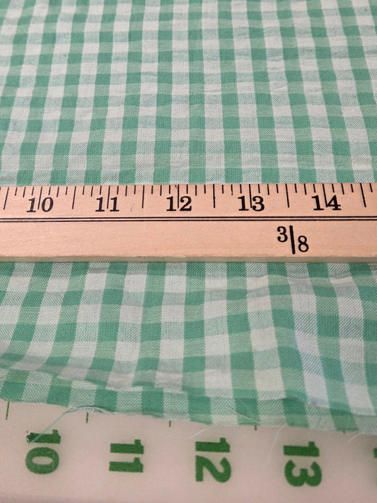 Green and white gingham checkered fabric shown with a wooden ruler for scale on a white cutting mat.