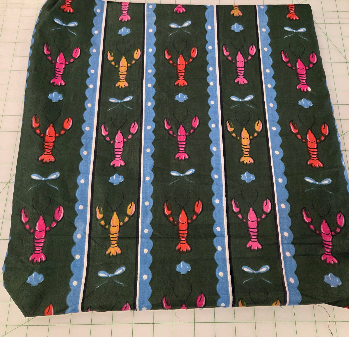 A square piece of fabric with a repeating lobster pattern on a dark green background, laid flat on a green grid cutting mat with a wooden ruler visible in the top right corner.