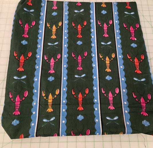 A square piece of fabric with a repeating lobster pattern on a dark green background, laid flat on a green grid cutting mat with a wooden ruler visible in the top right corner.