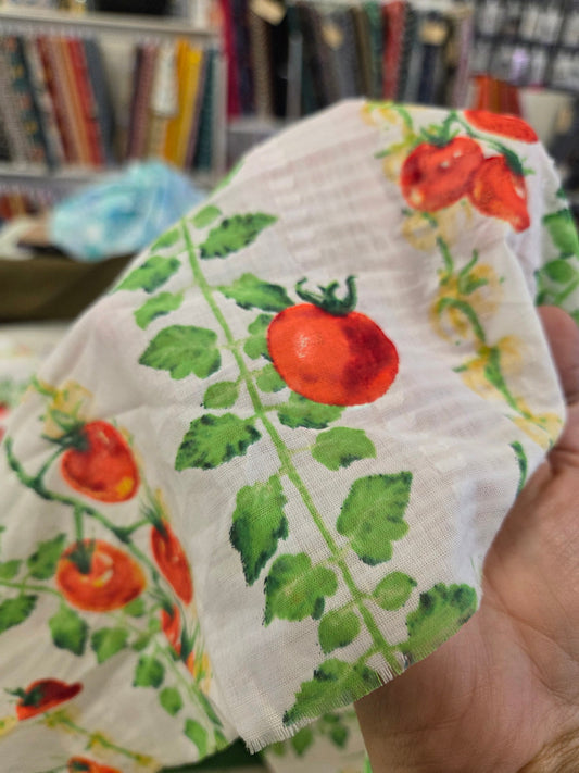 A close-up of a hand holding a white gauze fabric with a watercolor-style print of red tomatoes on green vines, with a blurred bookshelf in the background.