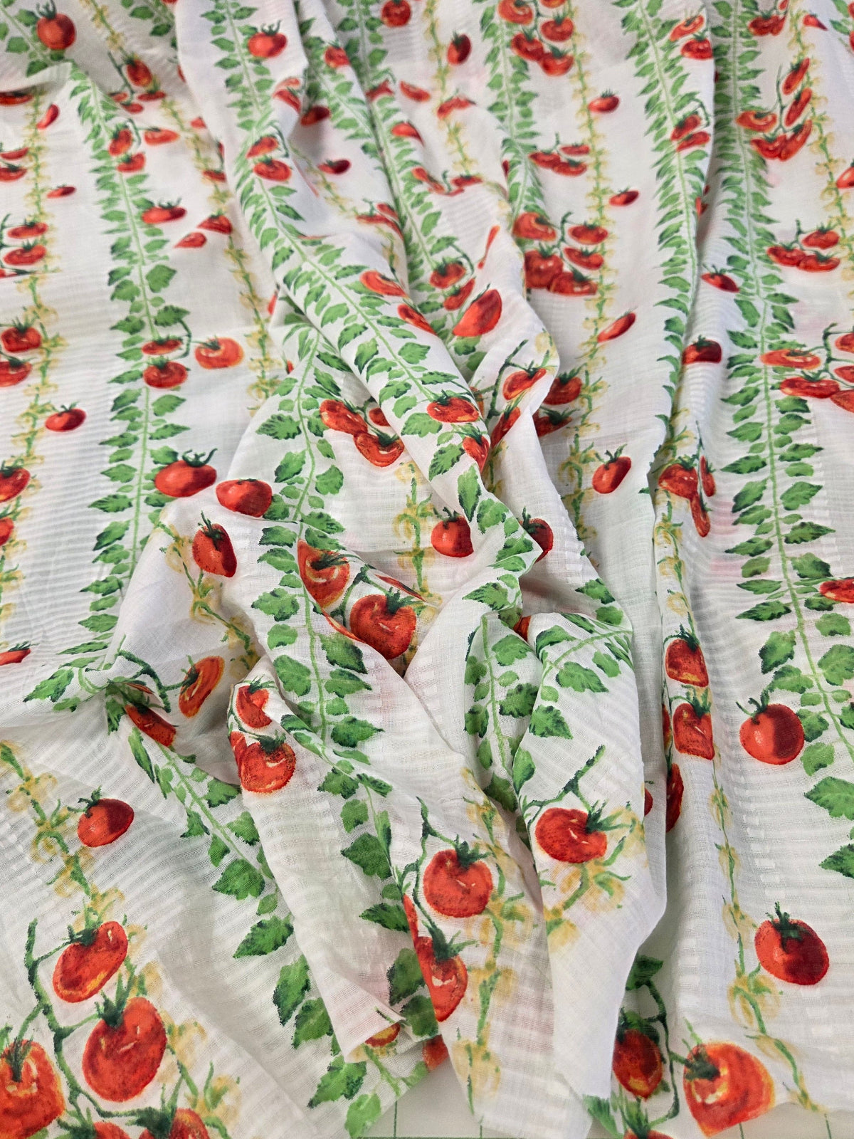 White crinkle gauze fabric printed with a repeating pattern of red tomatoes on green vines, draped to show its texture and print.