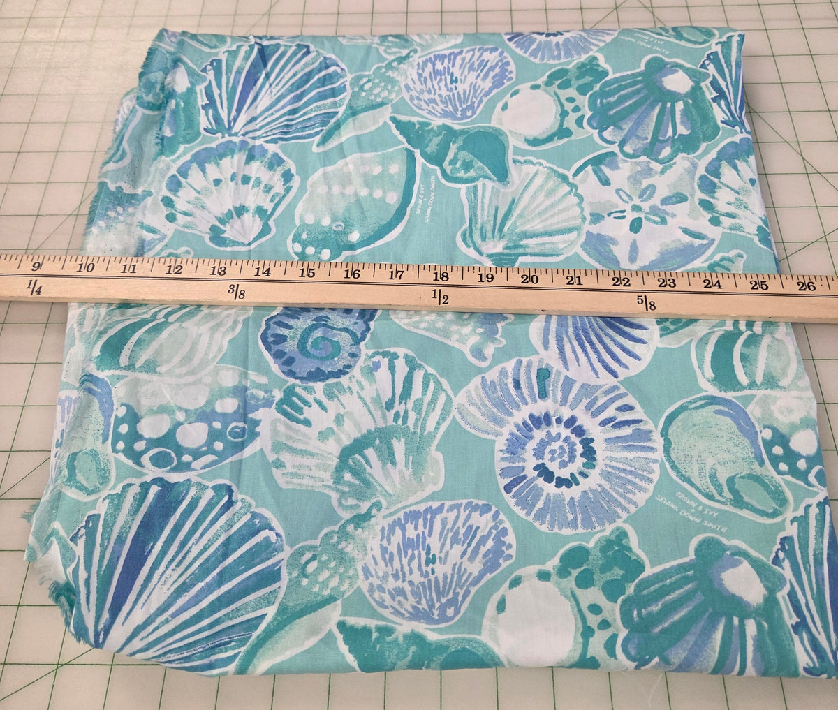 A folded piece of fabric with a blue and white seashell pattern on a mint green background, shown with a wooden ruler to indicate its width on a green grid cutting mat.