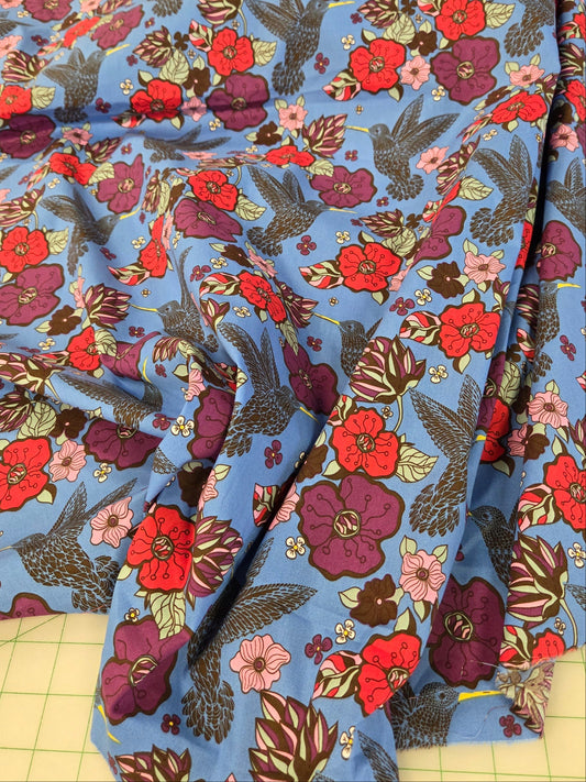A light blue fabric with a dense floral and botanical print in red, purple, pink, green, and brown, draped over a cutting mat.