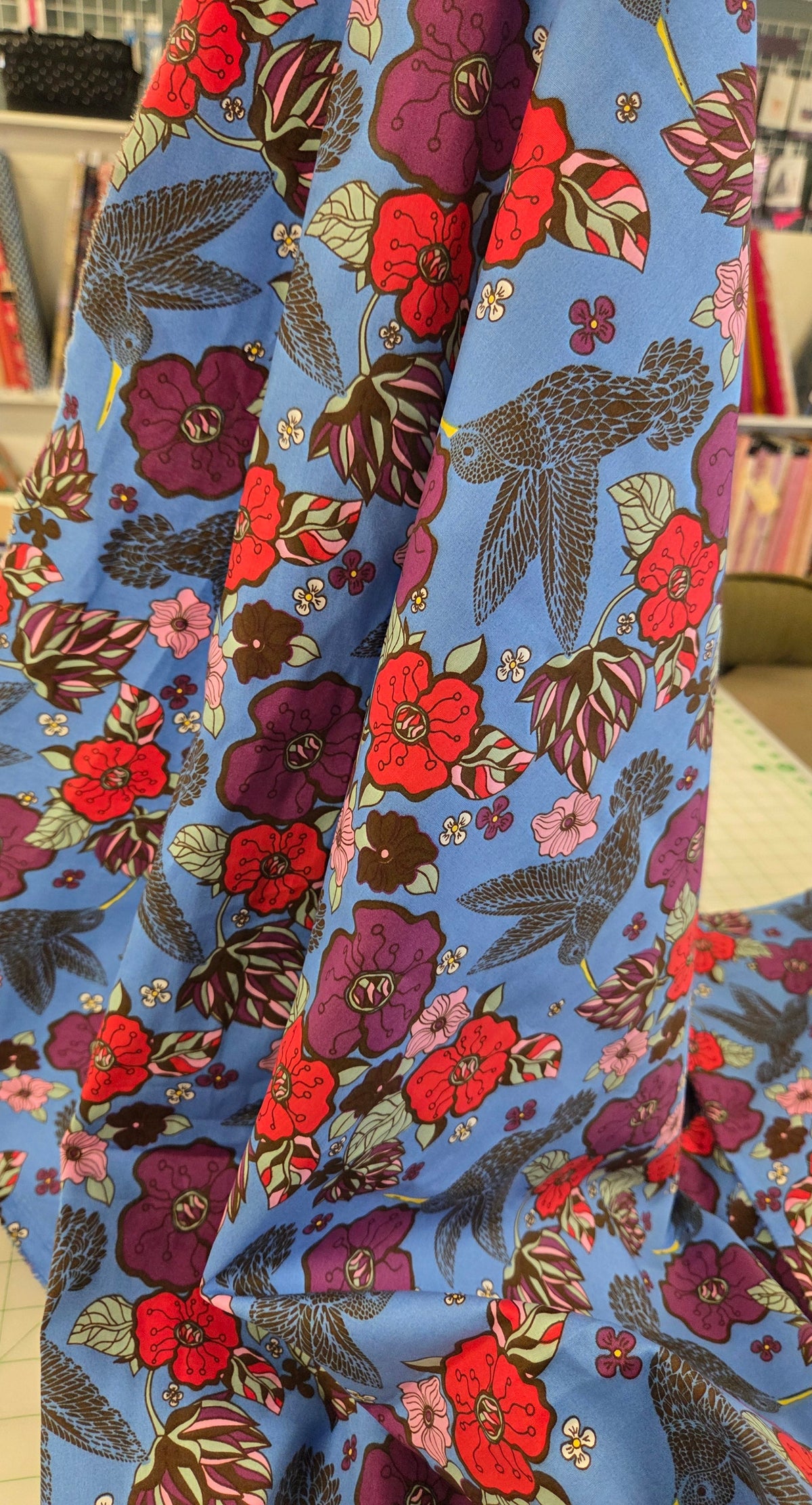 A close-up of a light blue fabric with a dense floral and bird print, draped over a table in a sewing or fabric store. The print features red, purple, and pink flowers with dark green and brown leaves and birds on a light blue background.