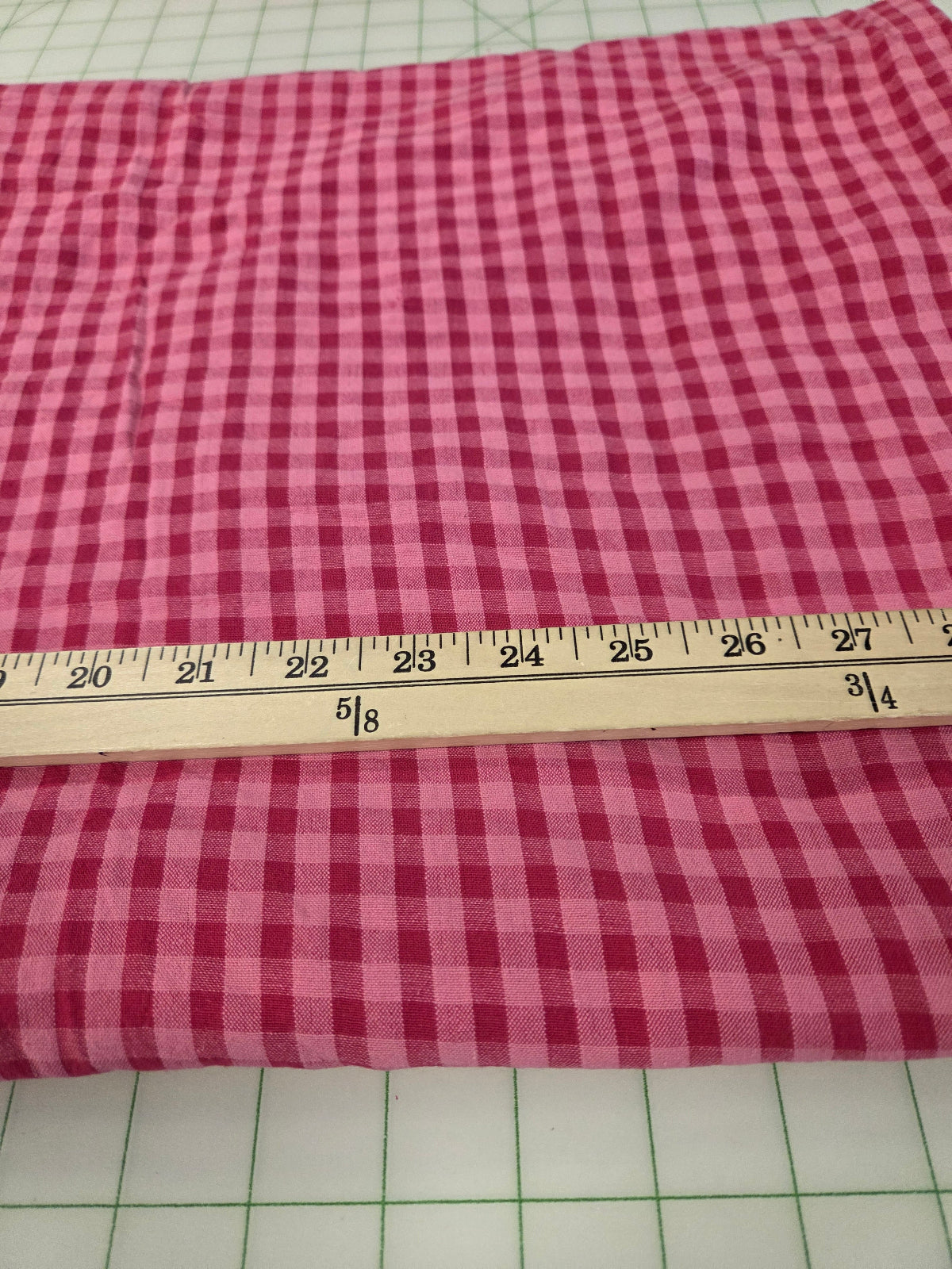 A folded piece of pink and red gingham fabric laid flat on a green-grid cutting mat, with a wooden ruler placed horizontally to show the pattern scale.