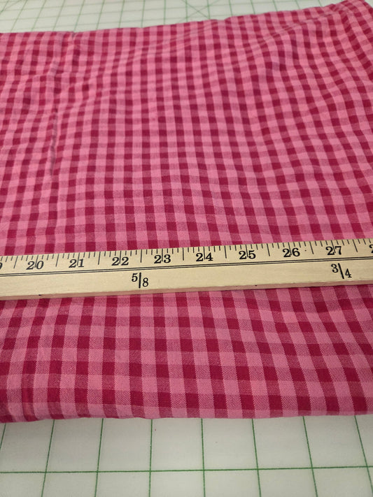 A folded piece of pink and red gingham fabric laid flat on a green-grid cutting mat, with a wooden ruler placed horizontally to show the pattern scale.