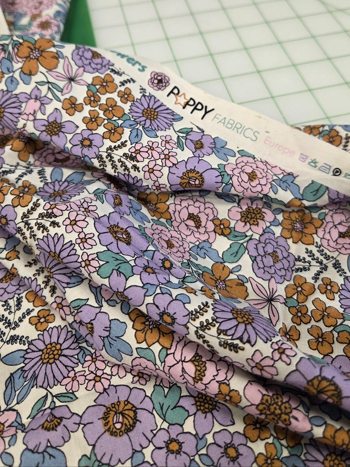 A close-up of floral fabric with a repeating print of purple, pink, orange, and green flowers on a cream background,