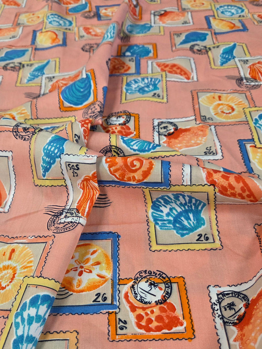 Pink fabric with a colorful seashell and shell-shaped postage stamp pattern, laid flat on a green cutting mat.