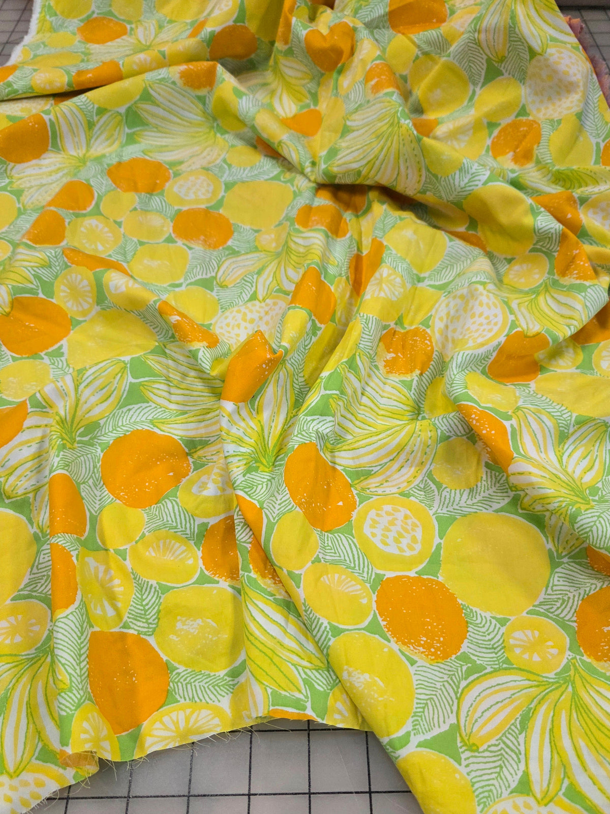 A piece of patterned fabric with a bright yellow and orange citrus fruit print on a light green background, laid out on a gridded cutting mat.