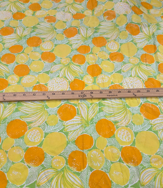 A piece of fabric with a vibrant citrus print featuring oranges, lemons, and limes on a light green background, laid flat on a cutting mat with a wooden ruler placed horizontally to show the width.