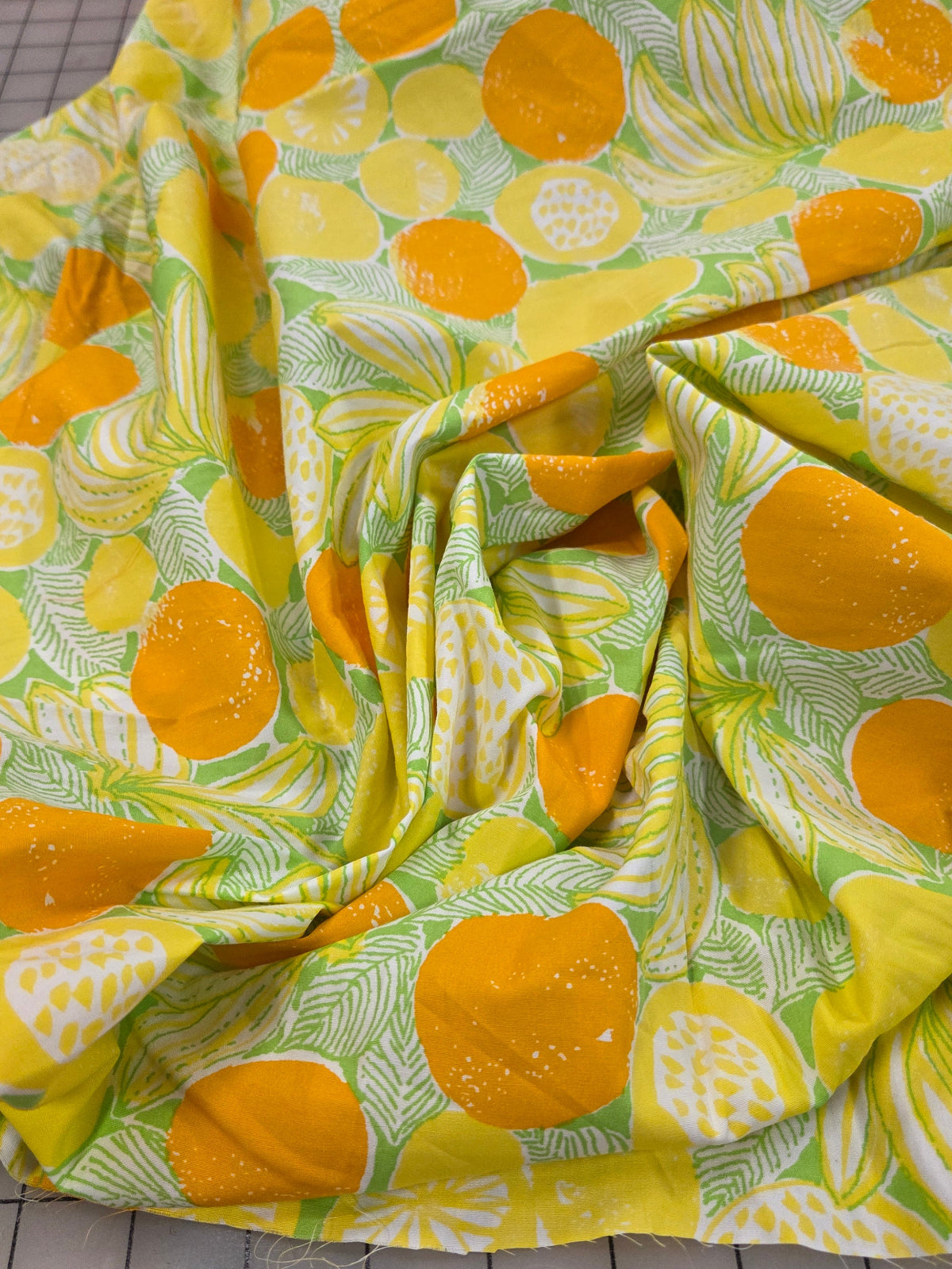 A close-up of a draped fabric with a vibrant citrus print featuring yellow and orange lemons and limes on a light green background with white leaf patterns, laid out on a gray grid cutting mat.