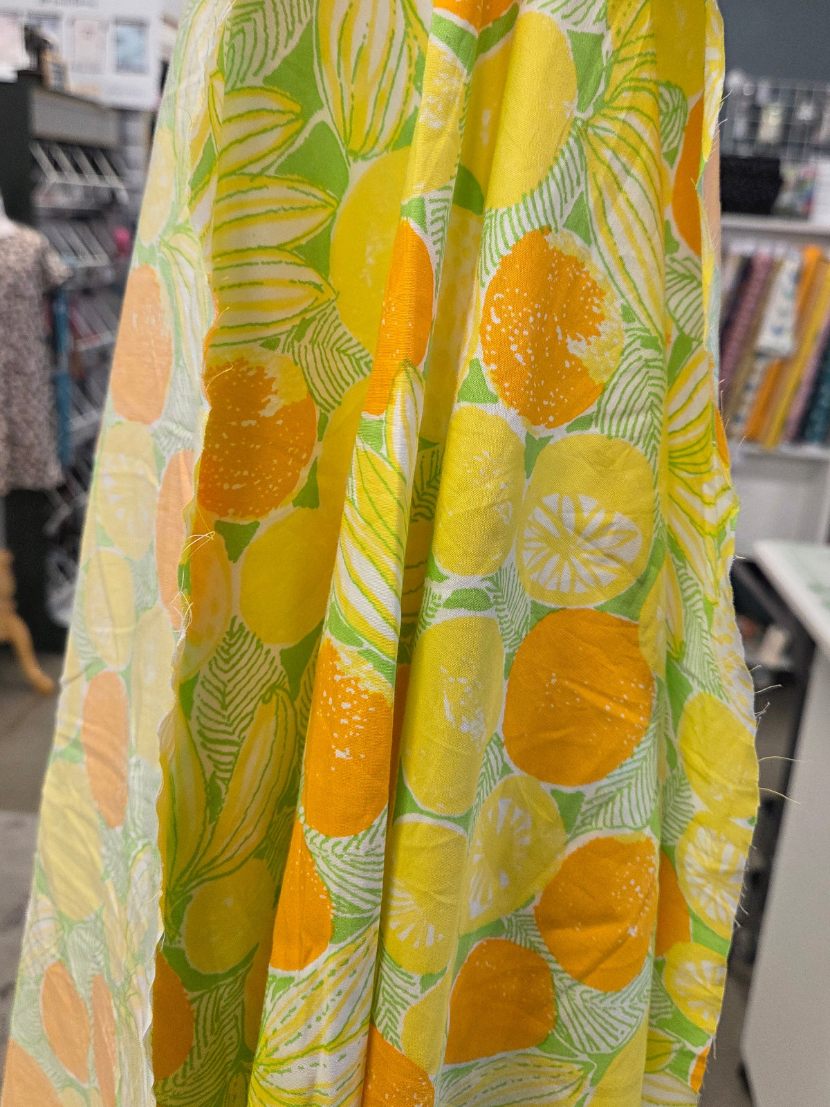 A close-up of a fabric or textile print featuring a bright yellow and orange citrus fruit pattern with green leaves on a white background, hanging in what appears to be a fabric store.