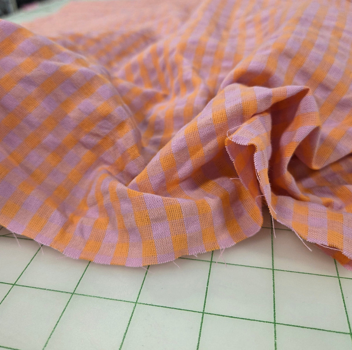 Pink and orange gingham check fabric draped over a white cutting mat with a green grid, with a blurred sewing or fabric shop background. LA Finch Fabrics designer deadstock  retail
