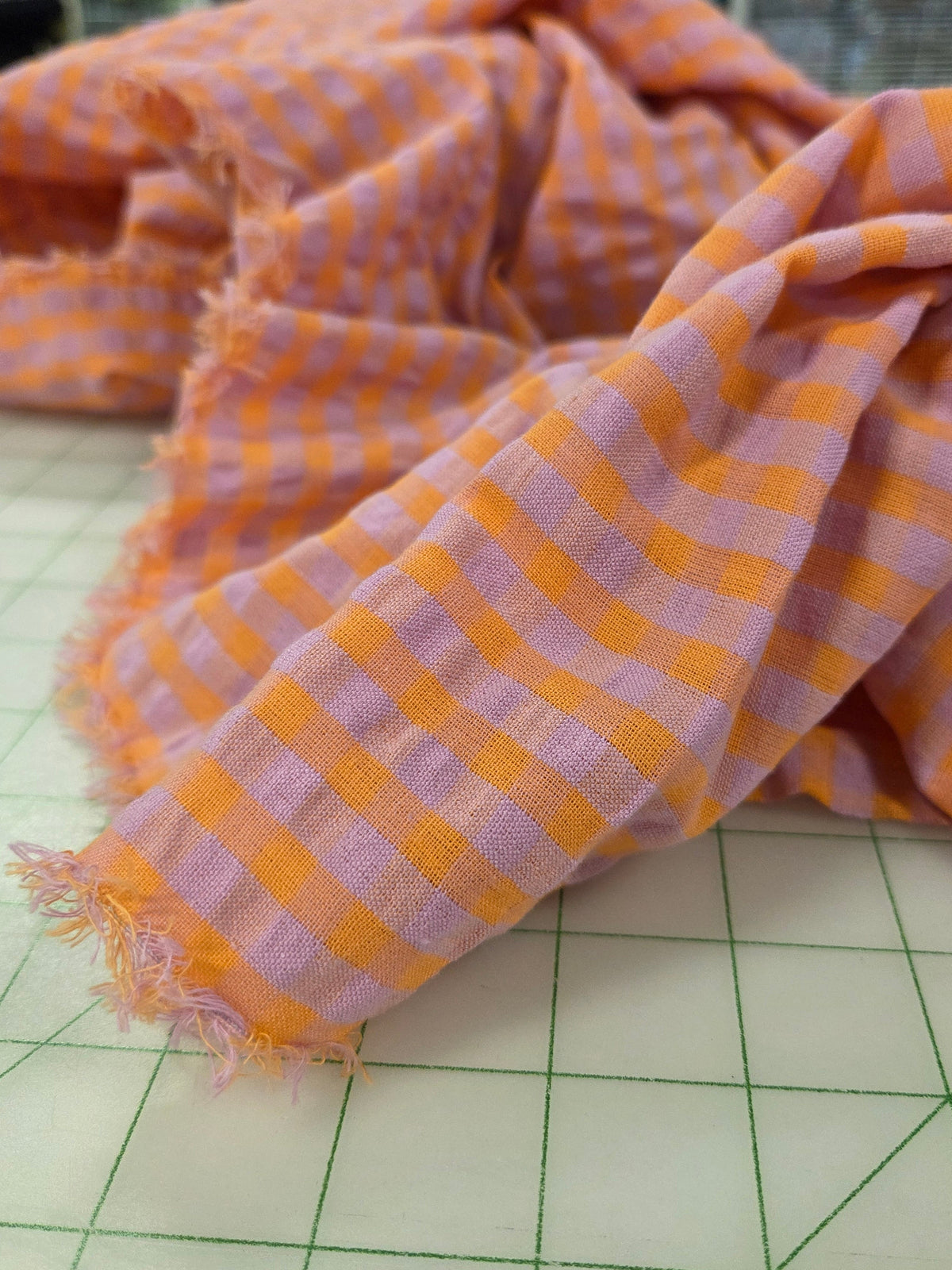 A close-up of a lightweight woven fabric with a pink and orange gingham check pattern and frayed edges, draped on a green-lined cutting mat.