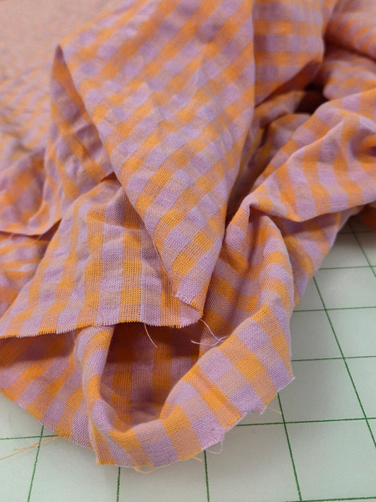 A close-up of draped fabric with a pink and orange gingham check pattern, laid on a white gridded cutting mat.