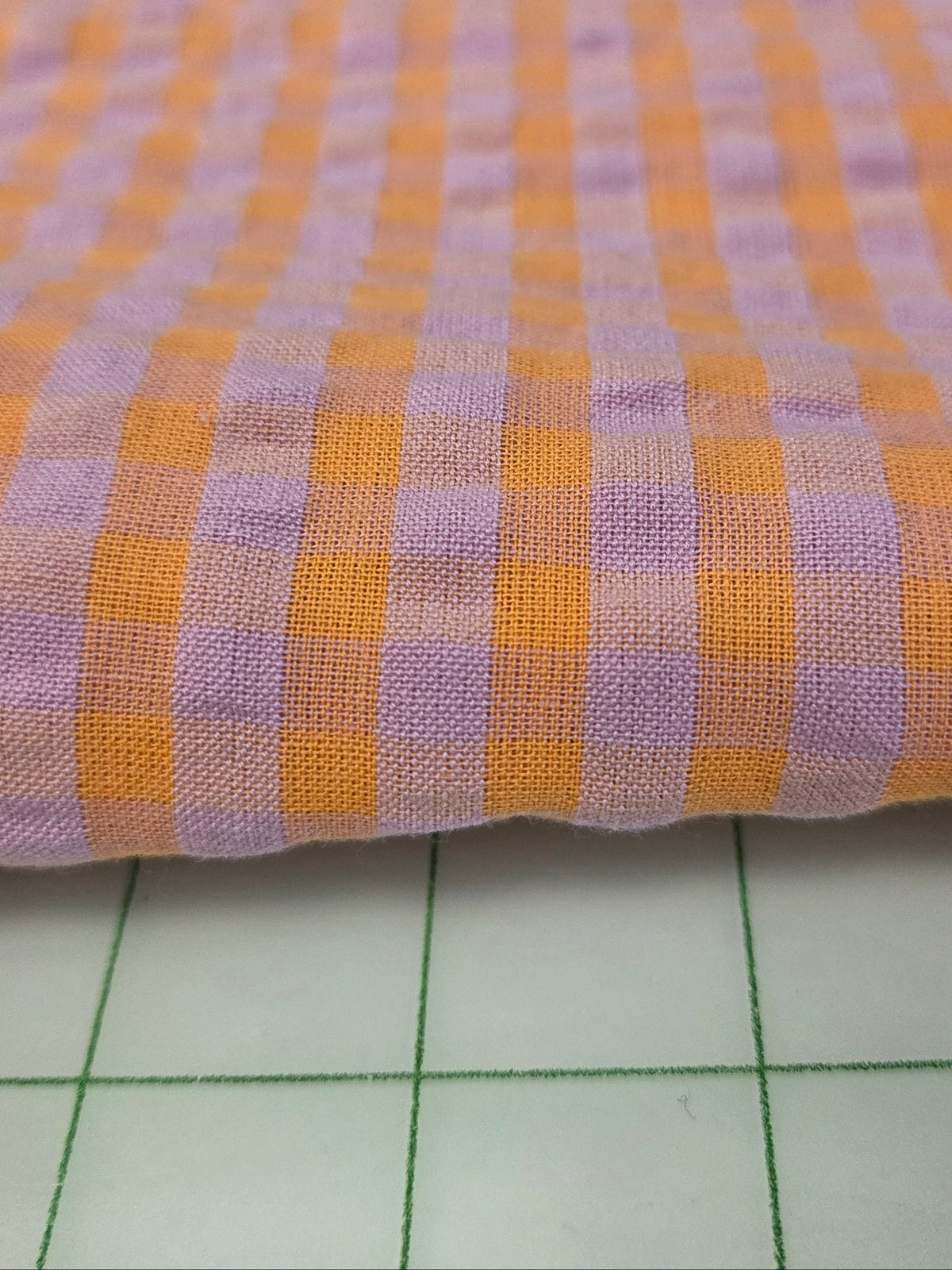 A close-up of a folded woven fabric with a gingham check pattern in orange and light purple, shown on a white cutting mat with a measuring tape visible at the top.