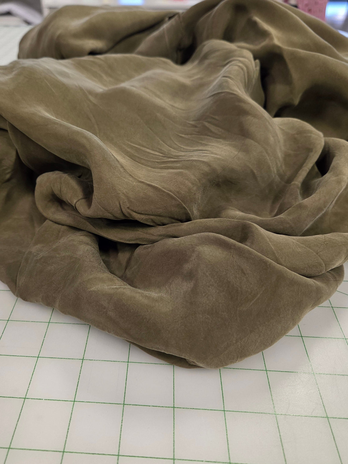 A crumpled olive green fabric piece, likely a scarf or textile swatch, resting on a white gridded cutting mat with a blurred craft room background.