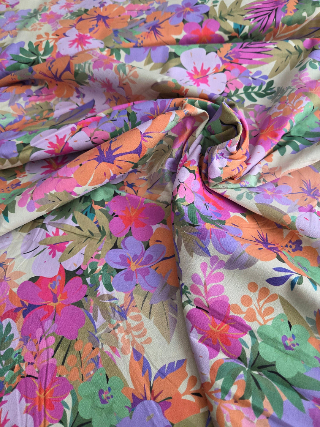 A close-up of a draped fabric with a dense, colorful floral pattern featuring pink, purple, orange, and green flowers and leaves on a light cream background.