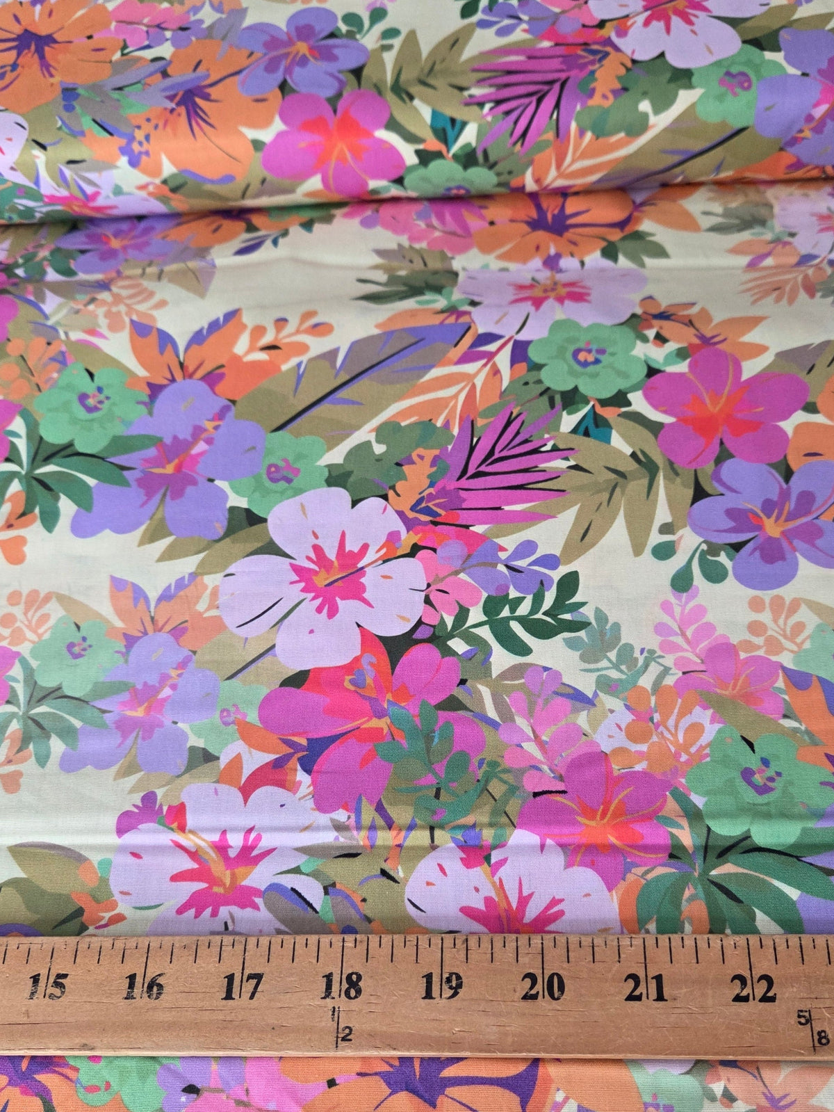 A close-up of a floral fabric with a multicolor tropical print, featuring pink, purple, orange, and green flowers on a light beige background. A wooden ruler is placed horizontally across the middle to show the scale of the pattern. LA Finch Fabrics