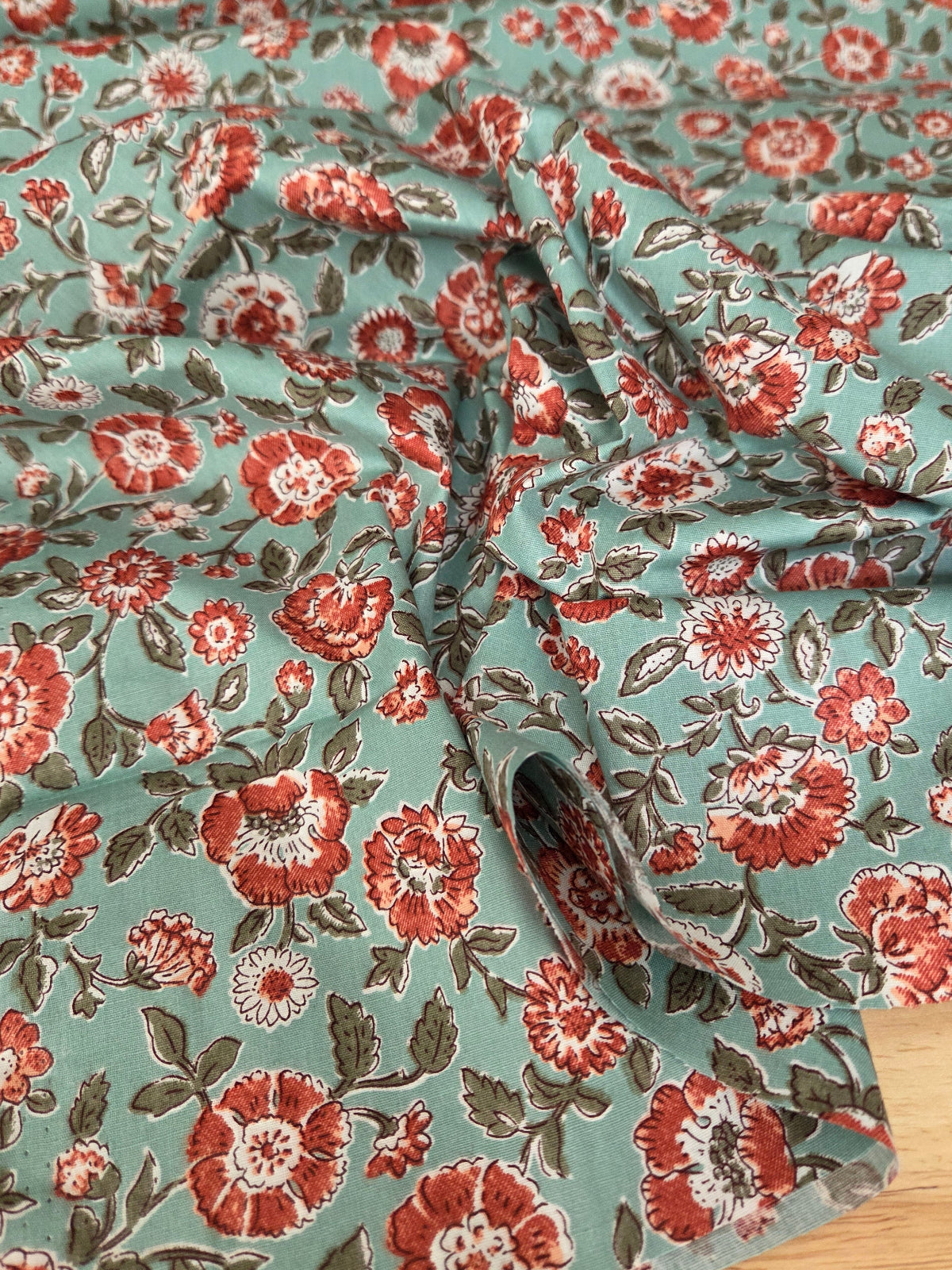 A close-up of draped fabric with a repeating floral block-print pattern on a muted sage green background, featuring red and orange flowers with white centers and dark green leaves, laid over a light wood surface.