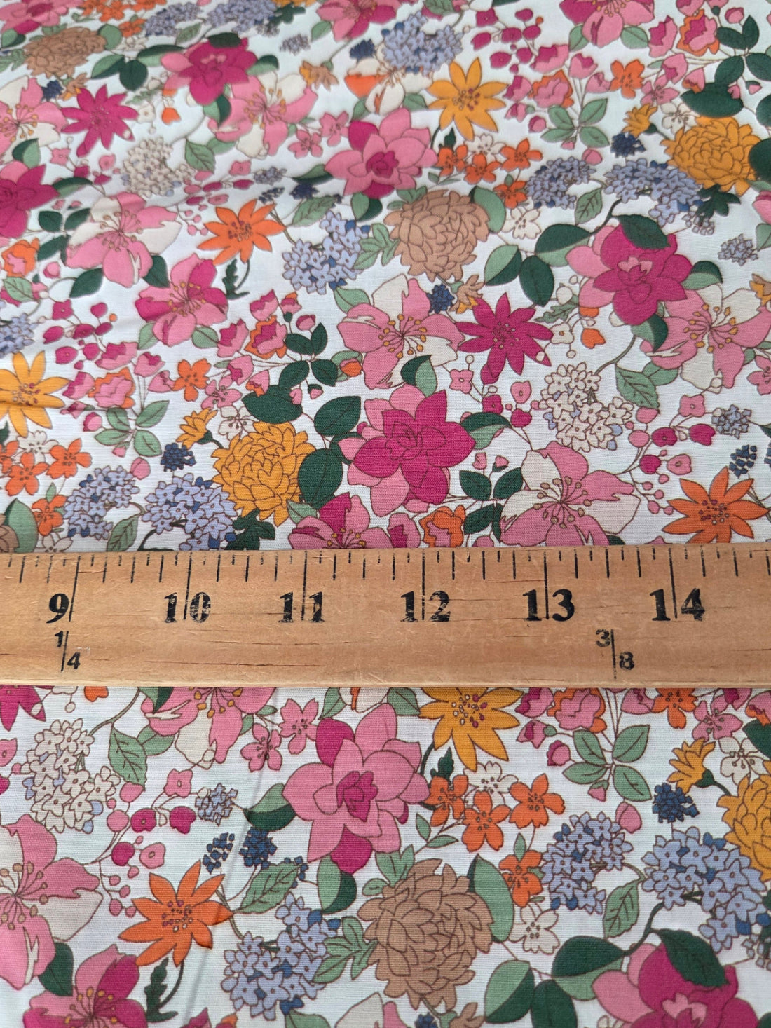 A close-up of floral fabric with a multicolor print of pink, orange, yellow, and purple flowers with green leaves on a white background, with a wooden ruler showing 15 inches placed horizontally to indicate scale.