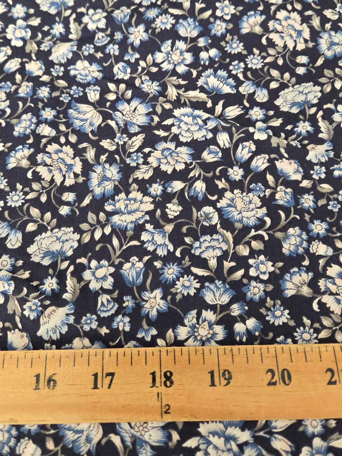 A close-up of a floral fabric with a navy blue background and light blue and white flowers, with a wooden ruler at the bottom showing the scale of the pattern.