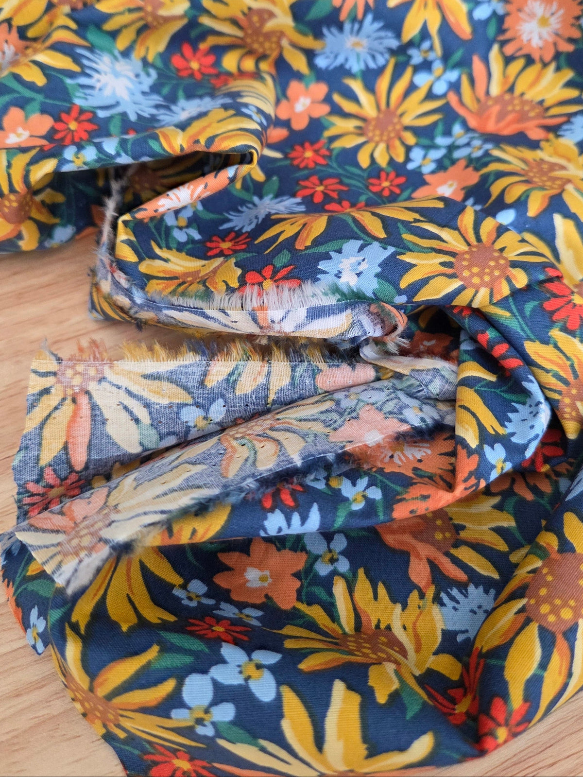 A close-up of a folded piece of fabric with a dense floral print in yellow, orange, blue, and white on a dark navy background, resting on a light wood surface.